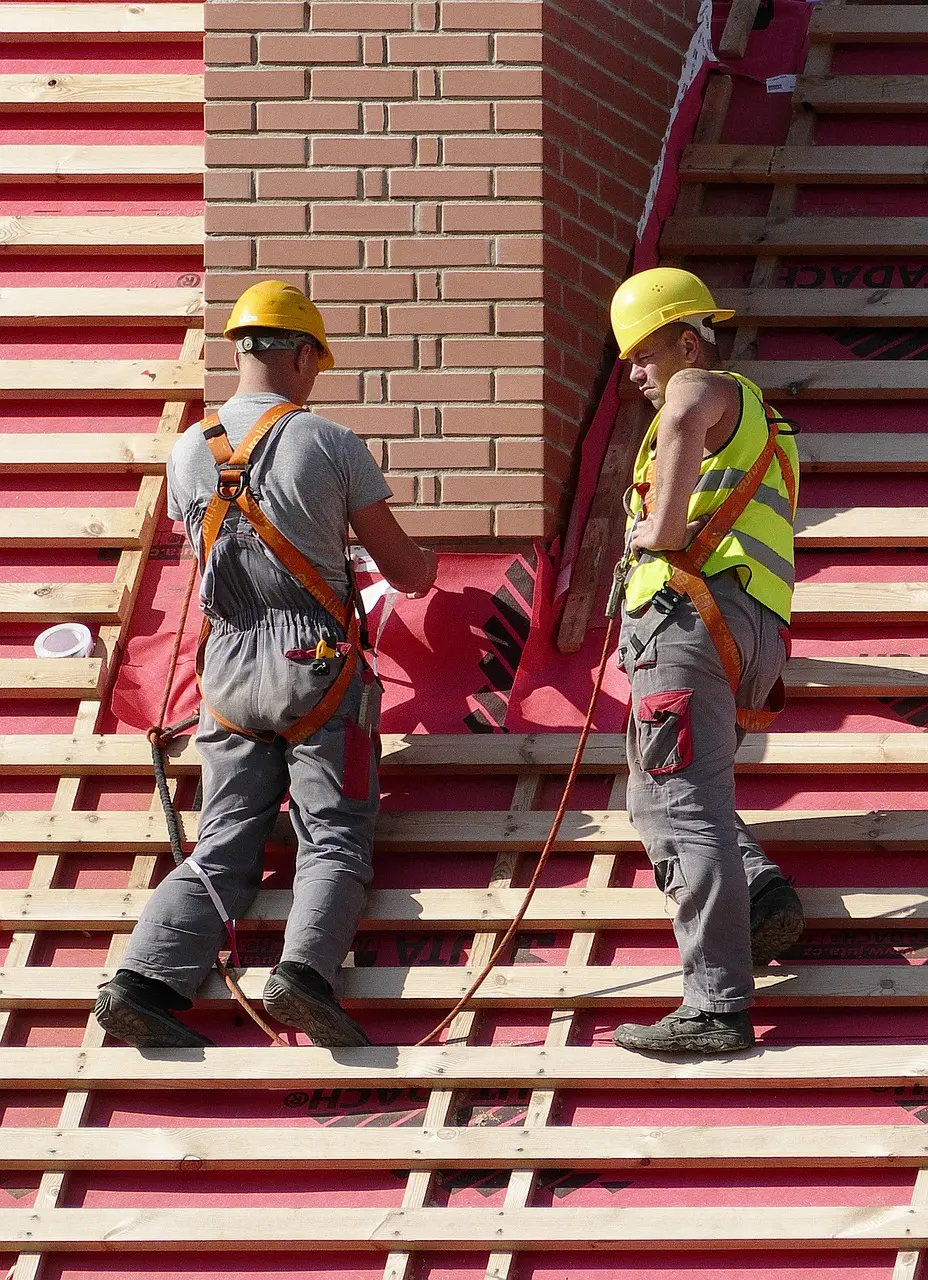 Two roofers in hard hats working on roof underlayment next to a chimney